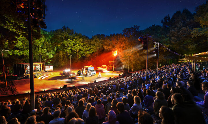 foto van een volle Bostheater tribune onder de sterrenhemel met de bomen en het podium verlicht met gele en rode lichten.