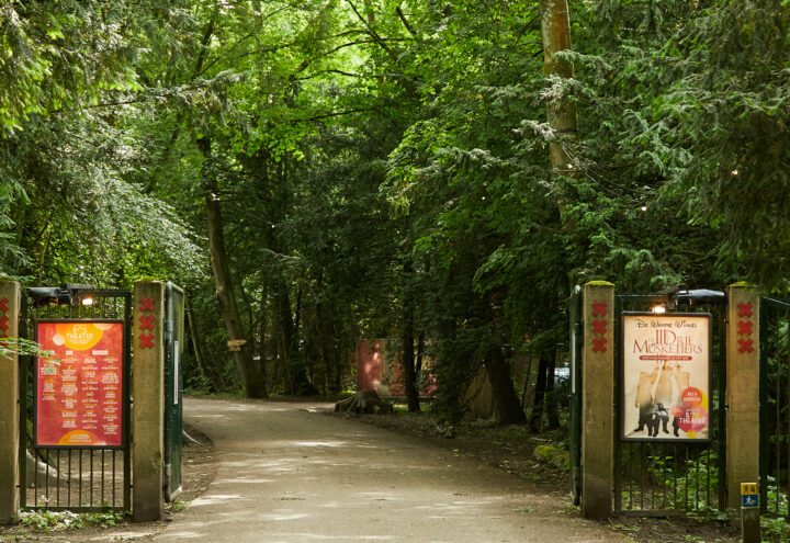 Foto van de ingang van het pad naar het Bostheater, met een hek dat open staat en aan weerszijde posters.